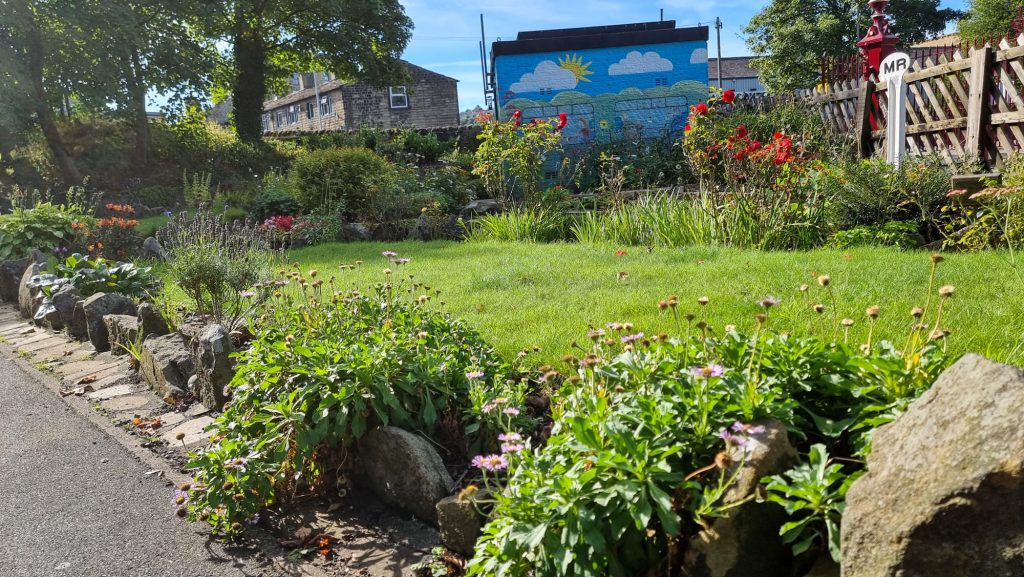 Image shows a lush garden. The border of the garden is lined with different shaped and sized stones, with plants growing freely over them. There is a grassed area with flower beds behind, and a building with a sunny sky murual painted on it.