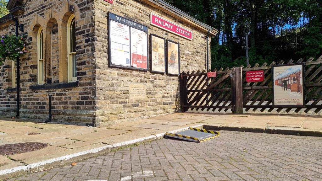 Image shows a wide angle shot of Ingrow West Station car park. The station building stands on the left, while the station fence and gate attaches to the building and follows to the right. In one disabled parking bay there is a small portable ramp to allow access to the pavement. The ramp is grey with yellow and black tape covers the sices and bottom lip. The ramp is wide enough for a wheelchair to alight.