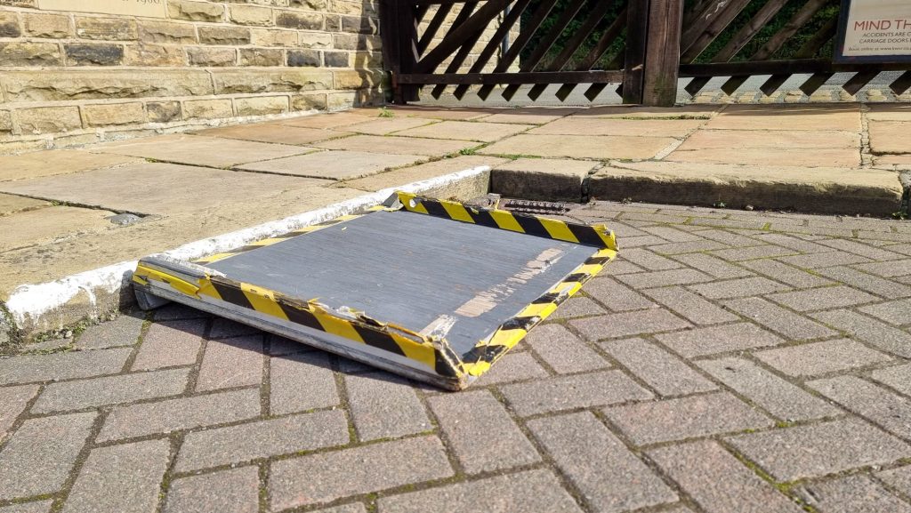 Image shows one disabled parking bay there is a small portable ramp to allow access to the pavement. The ramp is grey with yellow and black tape covers the sices and bottom lip. The ramp is wide enough for a wheelchair to alight.