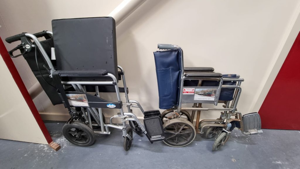 Image shows two folded wheelchairs against a wall in the Vintage Carriages Trust museum. These wheelchairs are for visitors to use if needed. One is larger than the other. Both have foot rests. Both chairs are to be pushed by someone, and cannot be self propelled.