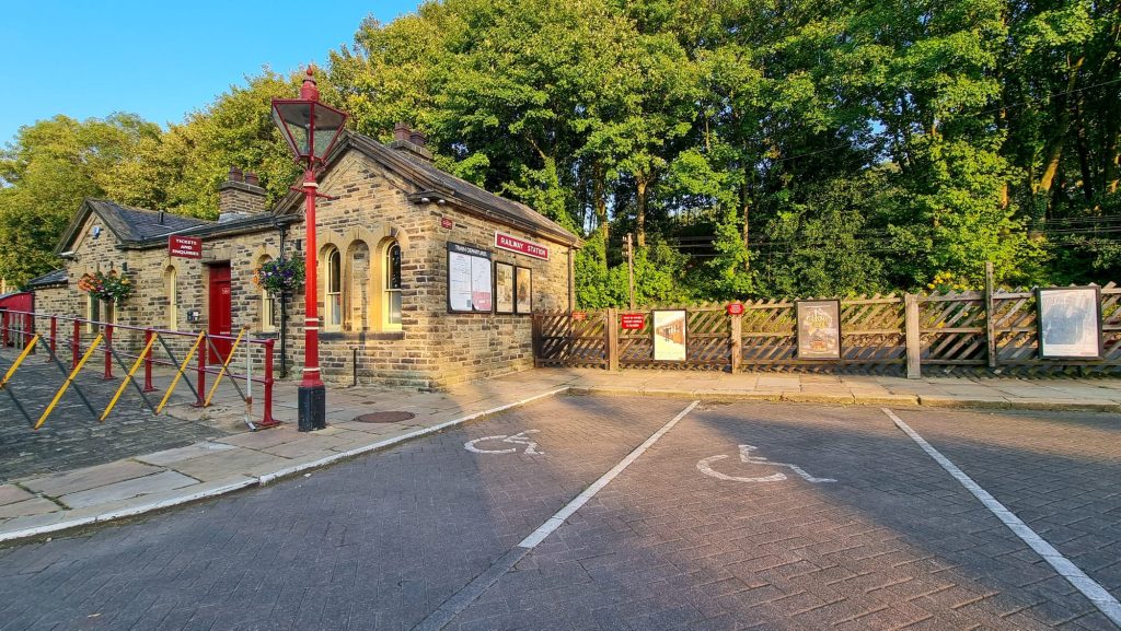 The image shows two disabled parking bays on the right hand side. Ingrow West Station is on the left of the image. There are green lush trees beside the station, and a clear blue sky shines in the top left of the image.