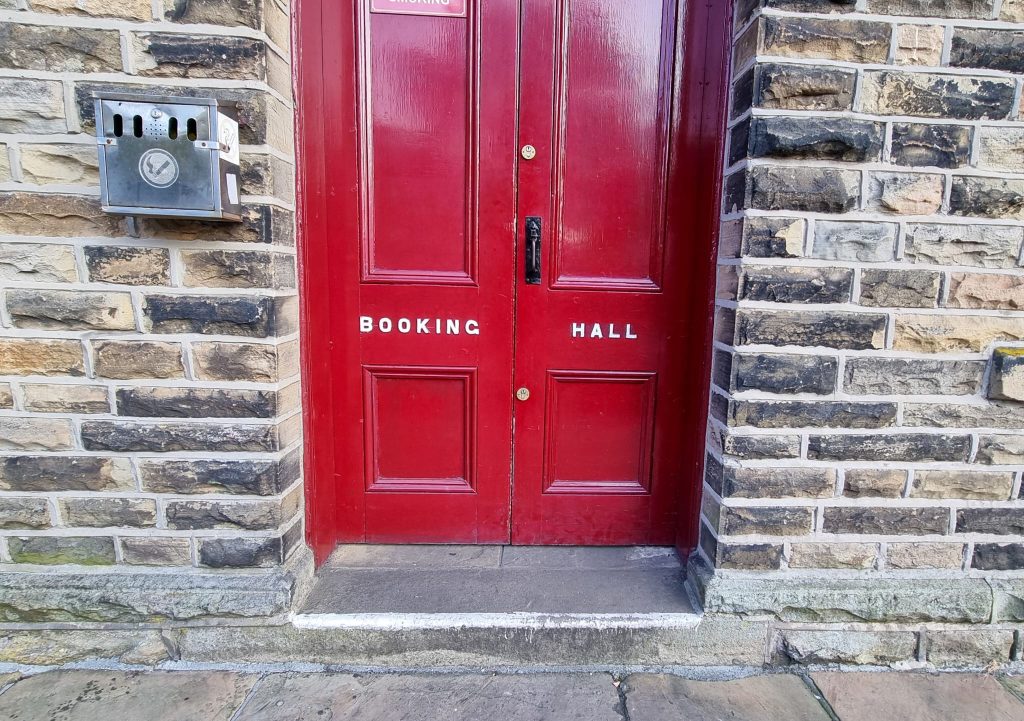 Image shows a red door which says 'Booking Hall' on it. There is a small step leading to the door of around 3.5 inches. The step is stone. There is a black door handle located on the right door in the middle.