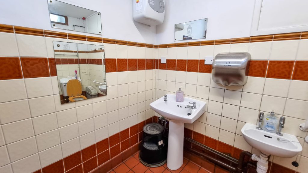 Image shows an accessibility toilet. From left to right there is a mirror, a foot stool beside a sink and a hand dryer.