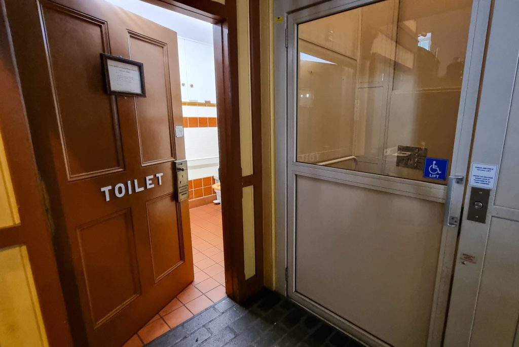 Image shows the toilet door slightly adjar. Next to the toilet door on the right is an accessibility lift.