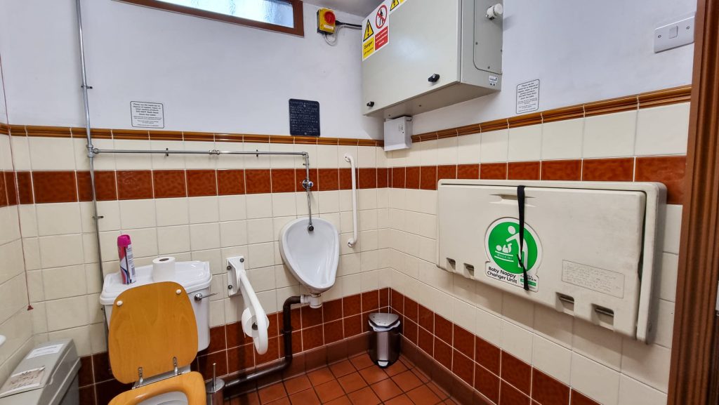 Image shows an accessibility bathroom. From left to right there is a toilet with hand rail, urinal with hand rail, a bin in the corner of the room, and a baby changing unit.