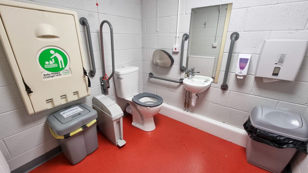 Image shows the accessible toilet in VCT. From left to right it shows a baby changing unit with a bin underneath. A toilet with support rails and an emergency pull chord, a sink and mirror with support rails and a paper towel dispenser with a bin underneath.